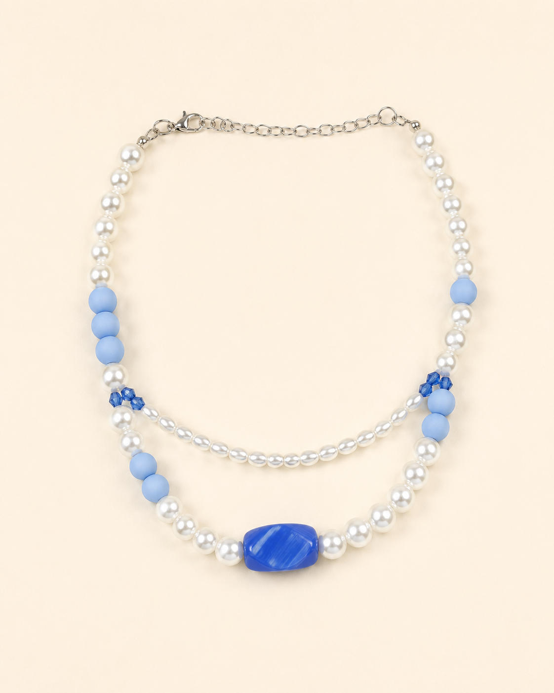 A double-strand beaded necklace featuring white faux pearls, light blue matte beads, a central blue oval stone, and small blue floral accents, displayed on a solid pale peach background.