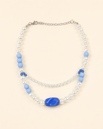 A double-strand beaded necklace featuring white faux pearls, light blue matte beads, a central blue oval stone, and small blue floral accents, displayed on a solid pale peach background.