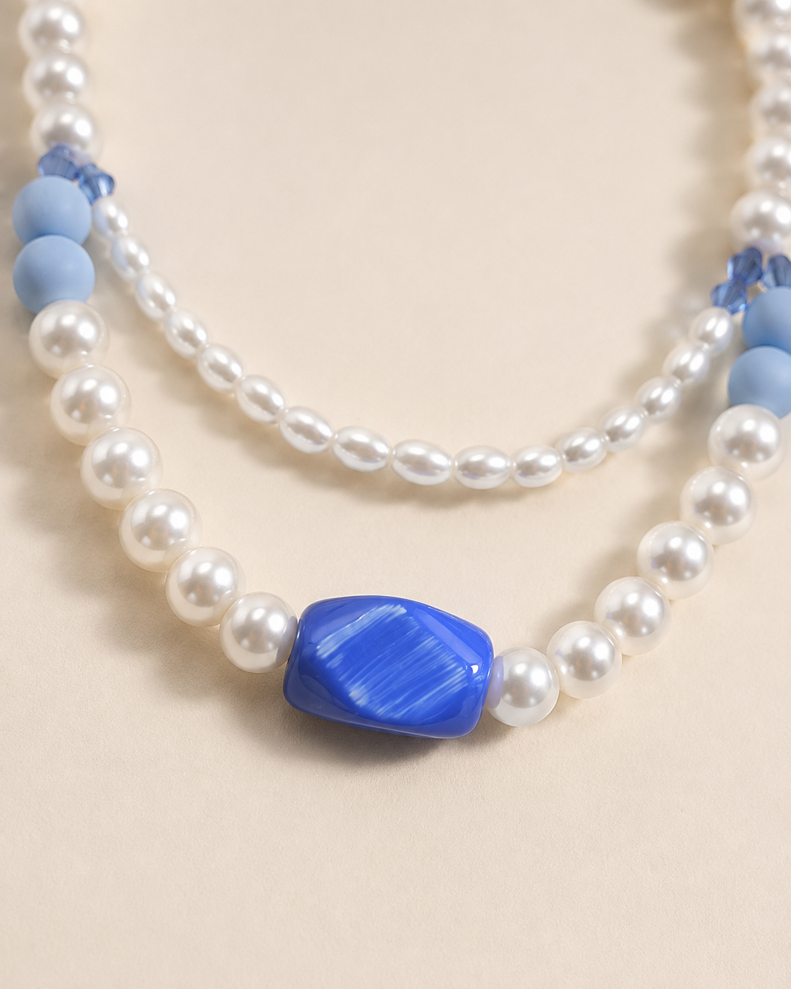 A double-strand beaded necklace featuring white faux pearls and smooth light blue beads, with a prominent textured blue oval centerpiece, displayed on a neutral beige background.