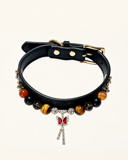 A black leather collar decorated with gold-tone hardware, polished brown and black beads, and a dangling silver bow charm embellished with red stones and clear crystals, set against a solid off-white background.