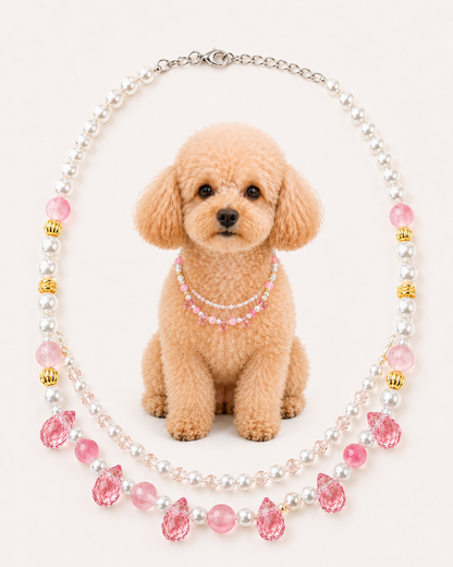A small tan poodle sits centered against a light cream background wearing a double-strand beaded necklace with white faux pearls, pink faceted beads, and small gold accents. The necklace is arranged in an oval around the dog, which appears to be modeling the product.