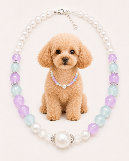 A small tan dog sits centered against a pale pink background wearing a short beaded collar necklace made of lavender and white beads. Around the dog is a larger pearl-style beaded necklace with alternating white pearls, light blue beads, and lavender beads separated by silver-toned spacer rings.