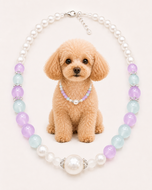 A small tan dog sits centered against a pale pink background wearing a short beaded collar necklace made of lavender and white beads. Around the dog is a larger pearl-style beaded necklace with alternating white pearls, light blue beads, and lavender beads separated by silver-toned spacer rings.