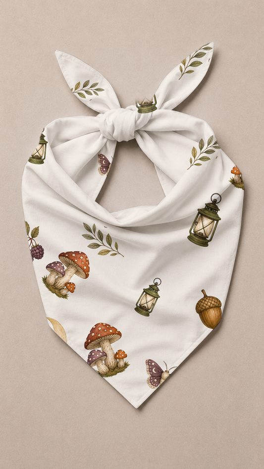 A white triangular bandana with a knotted bow, featuring an all-over print of mushrooms, lanterns, acorns, and leaves on a neutral beige background.