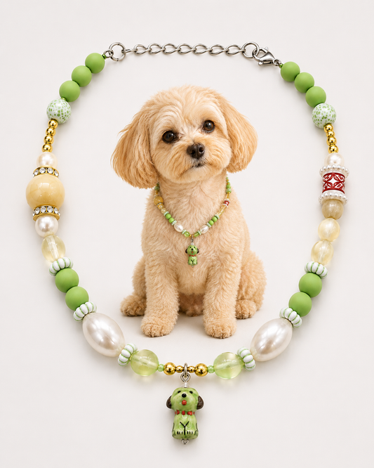 A small beige dog sits centered in front of a beaded pet collar necklace on a plain light background. The necklace features light green beads, white faux pearls, clear faceted beads, gold-toned spacer details, and a green dog-shaped charm hanging from a silver chain.