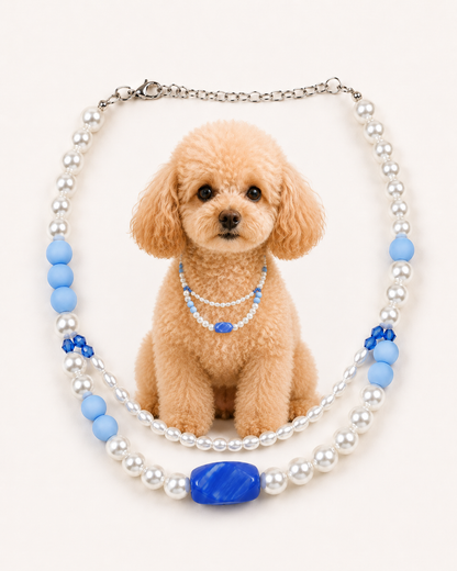 A decorative pet collar necklace featuring white faux pearls, light blue beads, and a large blue oval centerpiece, displayed around a small tan poodle on a plain light background.
