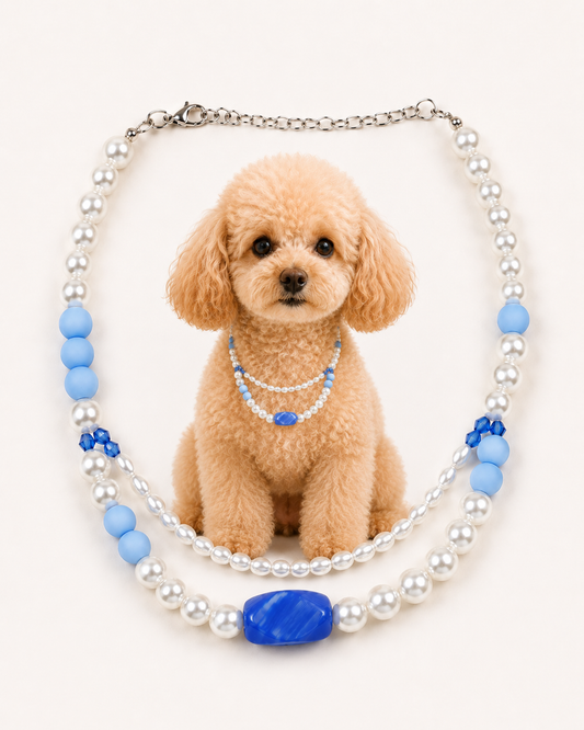 A decorative pet collar necklace featuring white faux pearls, light blue beads, and a large blue oval centerpiece, displayed around a small tan poodle on a plain light background.