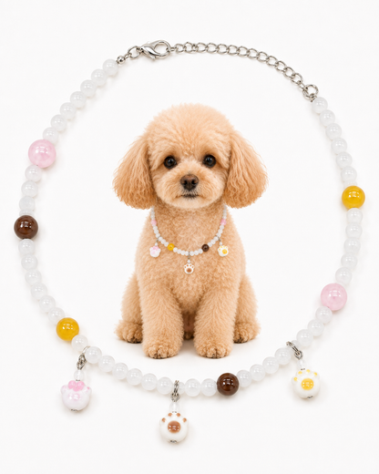 A small tan poodle sits centered in front of a beaded pet collar necklace on a white background. The necklace has translucent white beads, a silver chain, and colorful round beads and charms in pink, yellow, brown, and white with a paw-print detail.