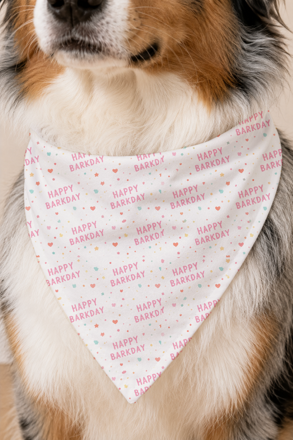 A close-up of a dog wearing a white triangular bandana with a repeating 'HAPPY BARKDAY' print in pink and light blue, with a fluffy brown, white, and black dog visible behind it.