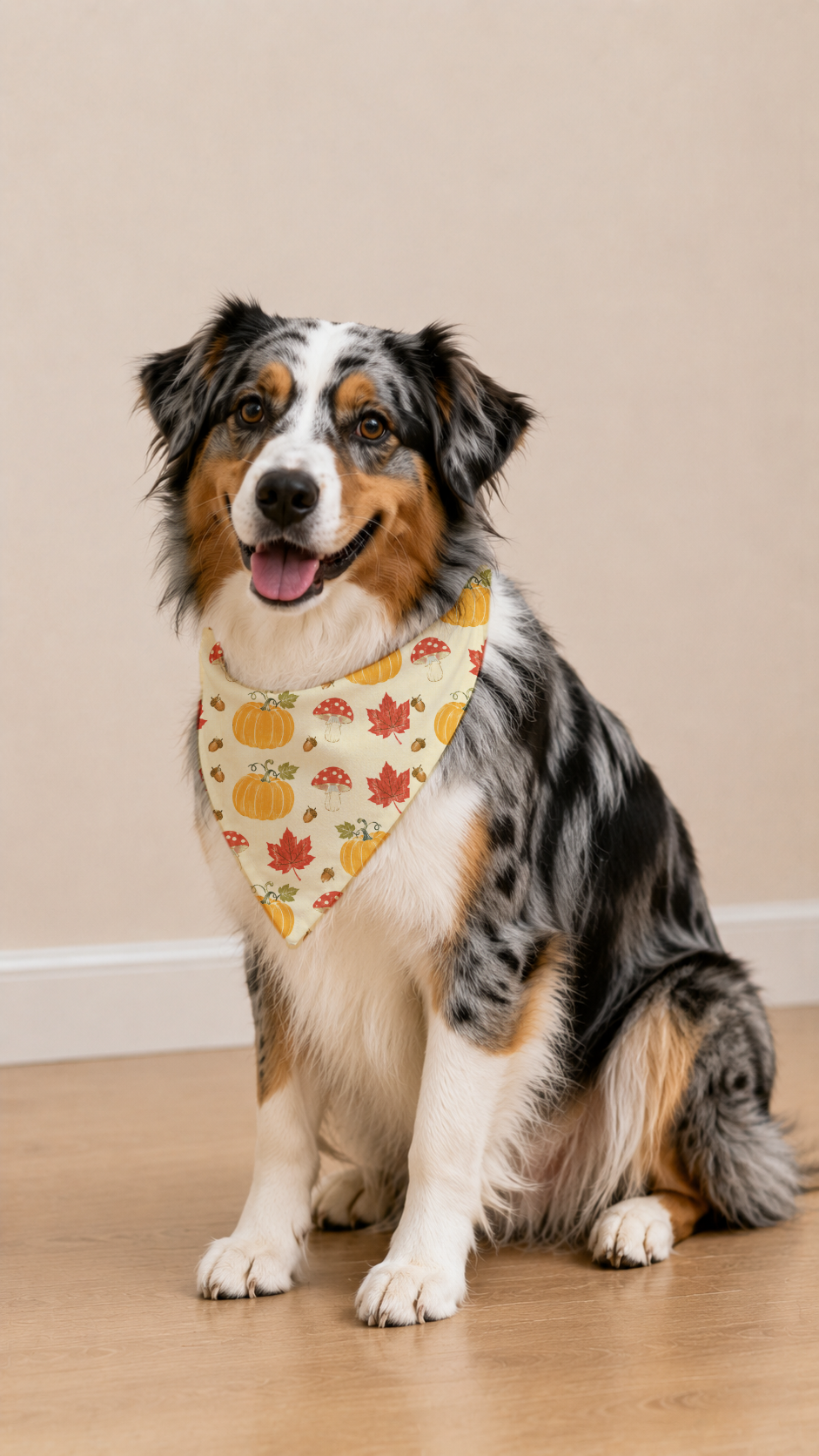 A long-haired Australian Shepherd dog sits on a light wood floor against a beige wall, wearing a cream bandana printed with orange pumpkins, red maple leaves, and small yellow flowers.