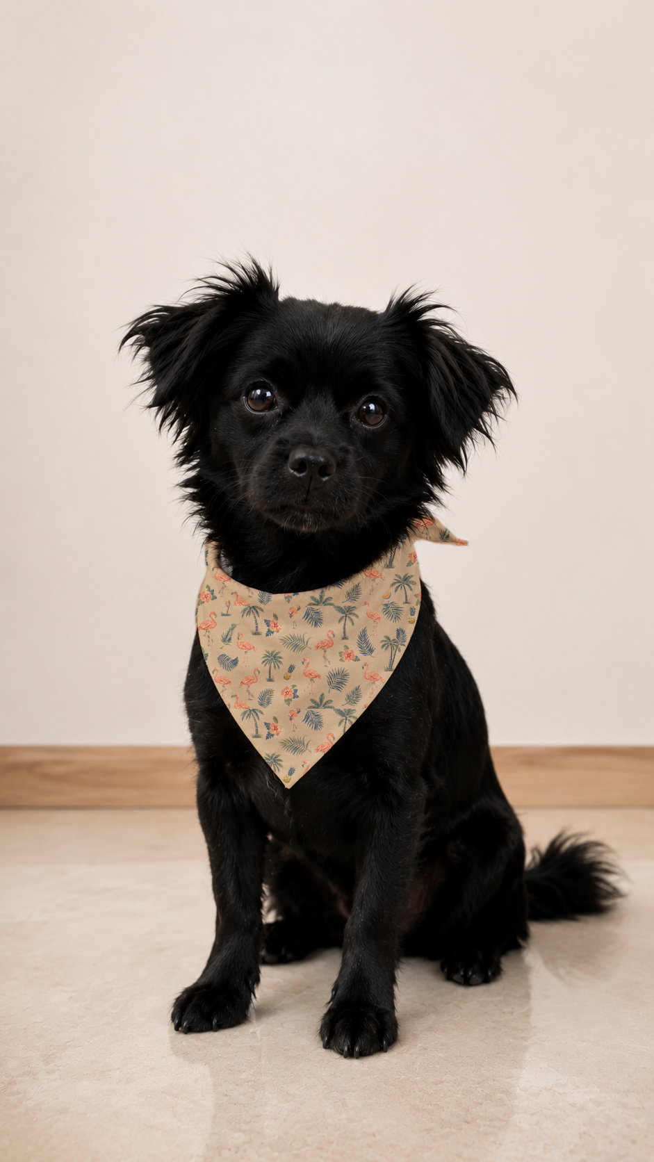 A small black dog wearing a beige bandana with a delicate floral pattern, sitting on a light beige floor against a neutral cream wall.