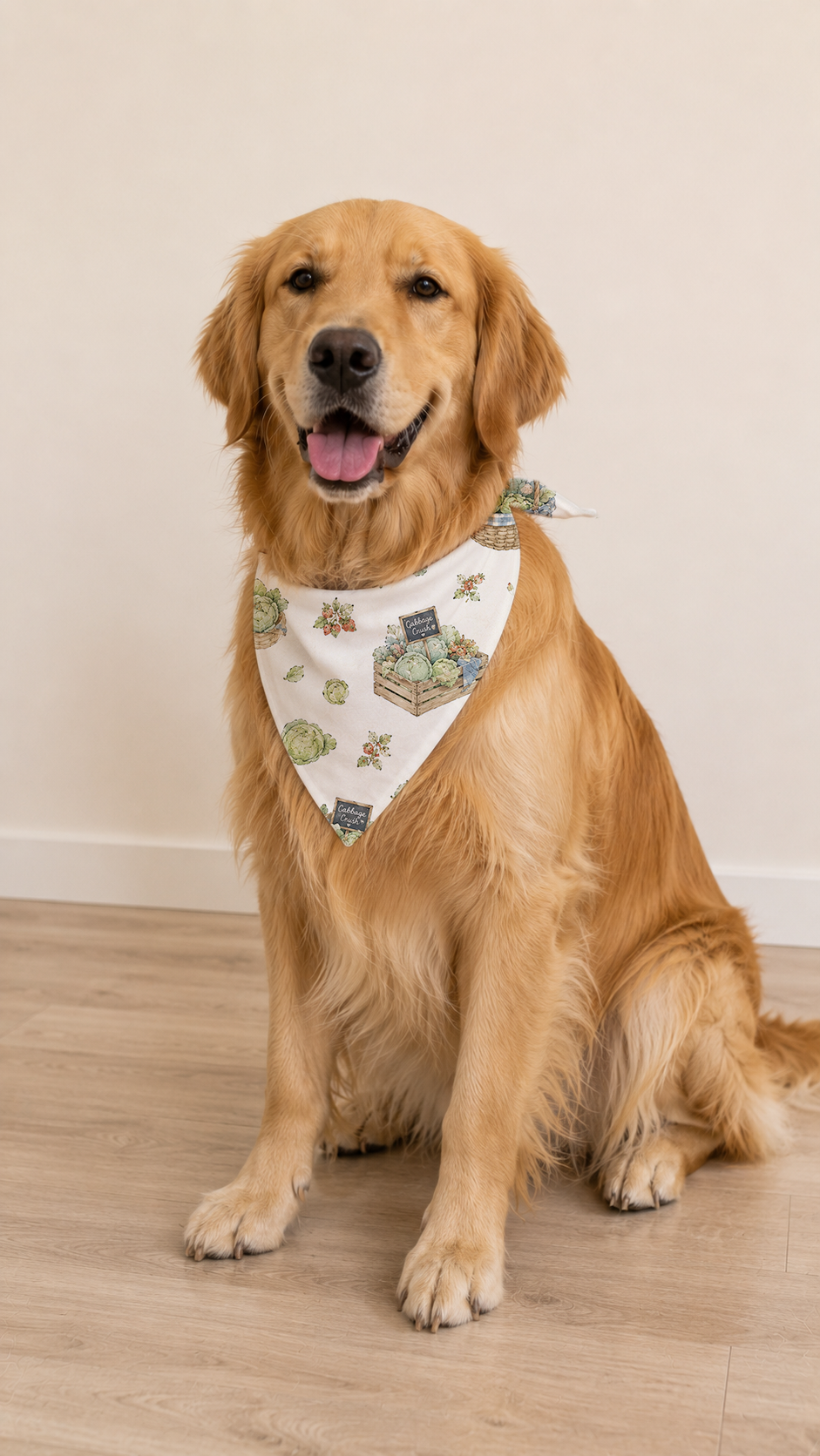 A golden retriever dog sitting on a light wood floor against a plain beige wall, wearing a white triangular bandana with a green and blue illustrated pattern.