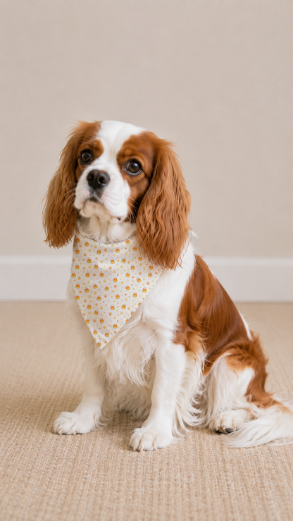 A small brown and white dog wearing a cream bandana with small yellow floral prints, sitting on a beige textured rug against a plain beige wall.