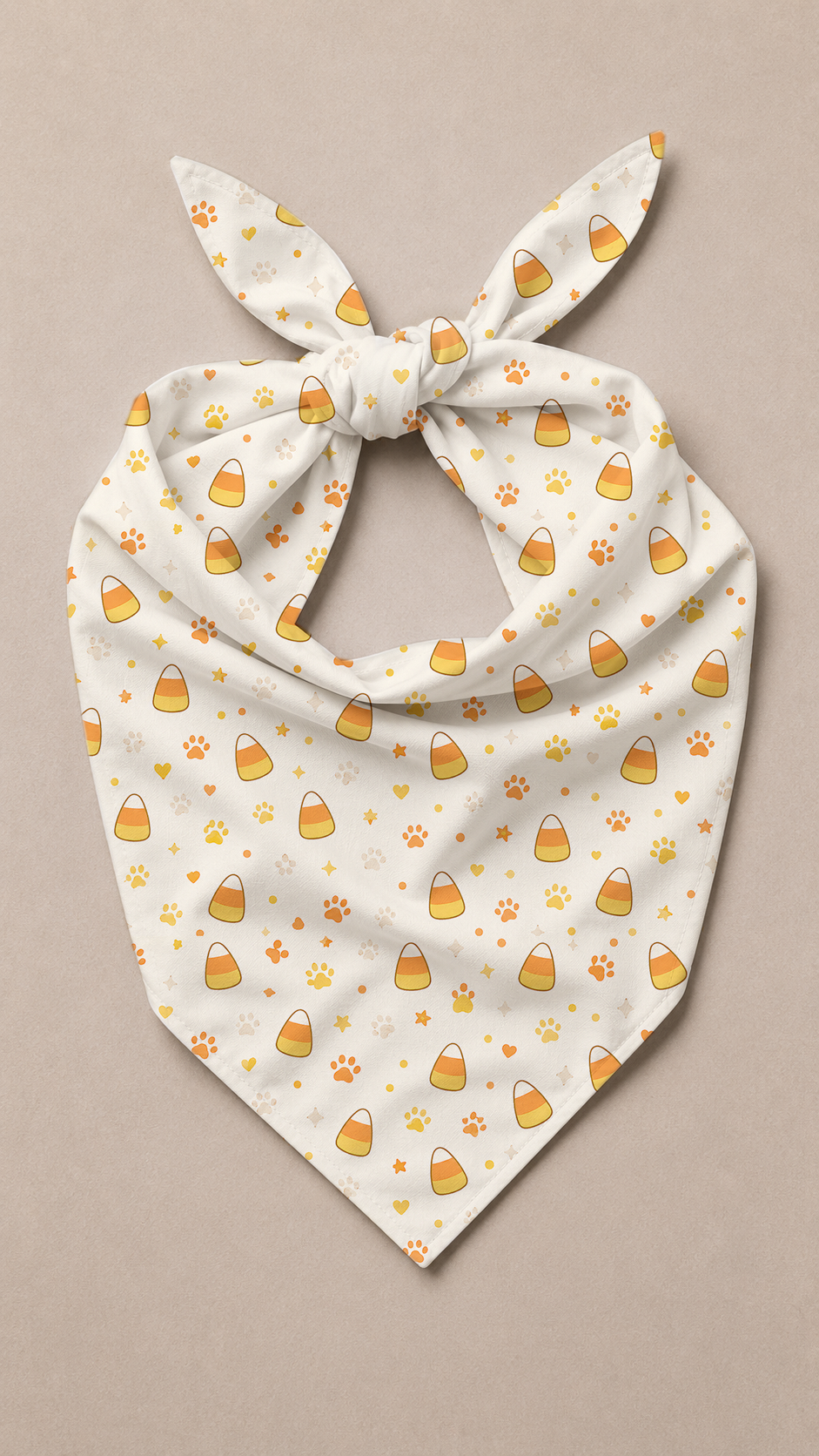 A cream-colored pet bandana with a tie knot, featuring a repeating pattern of yellow and orange candy corn and paw prints, displayed flat on a neutral beige background.