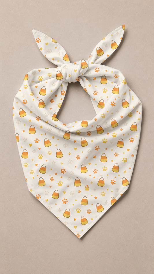 A cream-colored pet bandana with a tie knot, featuring a repeating pattern of yellow and orange candy corn and paw prints, displayed flat on a neutral beige background.