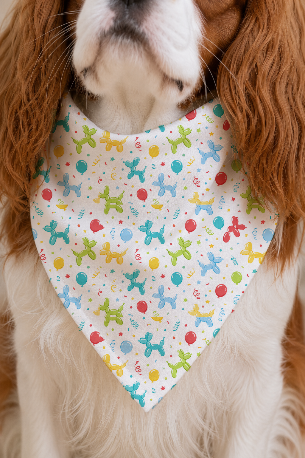 A close-up of a dog wearing a white triangular bandana bib with a colorful balloon-dog pattern, set against the dog's brown and white fur.