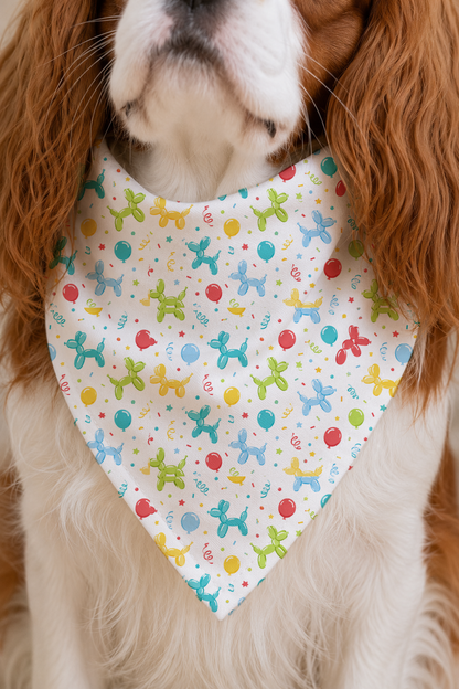 A close-up of a dog wearing a white triangular bandana bib with a colorful balloon-dog pattern, set against the dog's brown and white fur.