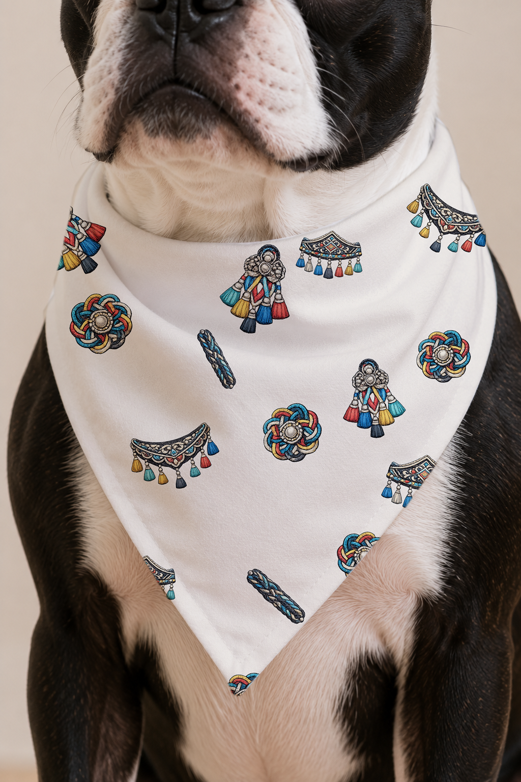 A close-up of a Boston Terrier dog wearing a white bandana printed with colorful traditional-style motifs, including figures, circular patterns, and tassel details.