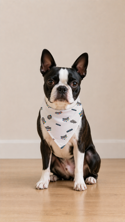 A Boston Terrier dog wearing a white bandana with a black cat print, sitting on a light wooden floor against a beige wall.