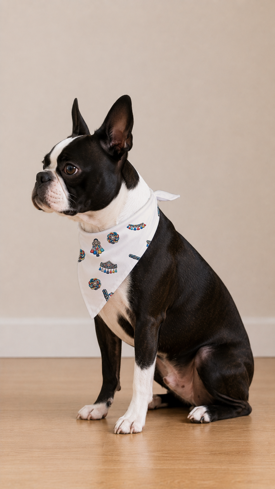 A Boston Terrier dog wearing a white bandana with a small blue and black pattern, sitting on a light wood floor against a beige wall.