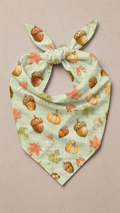 A light mint green pet bandana with a tie knot, featuring an all-over autumn print of pumpkins, acorns, and maple leaves, laid flat on a neutral beige background.