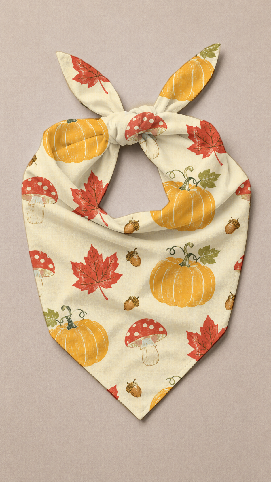 A cream-colored pet bandana with a tie-neck design, featuring an autumn pattern of orange pumpkins, red maple leaves, red-and-white mushrooms, and acorns, displayed on a neutral beige background.