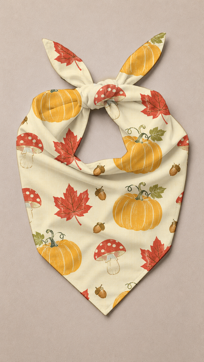 A cream-colored pet bandana with a tie-neck design, featuring an autumn pattern of orange pumpkins, red maple leaves, red-and-white mushrooms, and acorns, displayed on a neutral beige background.