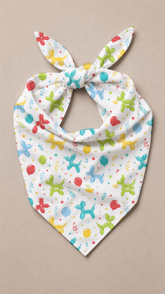 A white pet bandana with a tie-neck design, featuring a colorful pattern of balloon animals, balloons, and confetti, laid flat on a neutral beige background.