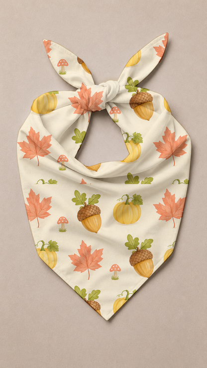 A cream-colored pet bandana with a tie-neck design, featuring an autumn pattern of orange maple leaves, acorns, pumpkins, and small mushrooms, laid flat on a neutral beige background.
