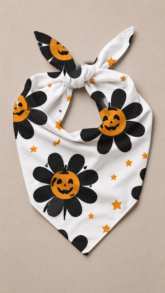 A white tie-on bandana or pet scarf featuring a black daisy pattern with orange pumpkin centers and small orange stars, laid flat on a beige background.