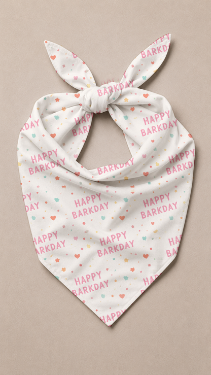 A white triangular pet bandana with a tie knot, featuring a repeating pattern of 'HAPPY BARKDAY' text in pink and small hearts in pink, orange, and mint green, displayed on a neutral beige background.
