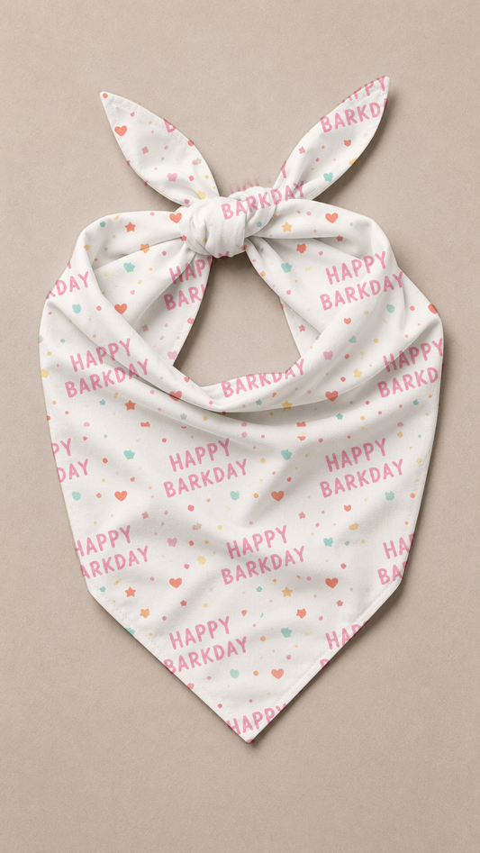 A white triangular pet bandana with a tie knot, featuring a repeating pattern of 'HAPPY BARKDAY' text in pink and small hearts in pink, orange, and mint green, displayed on a neutral beige background.