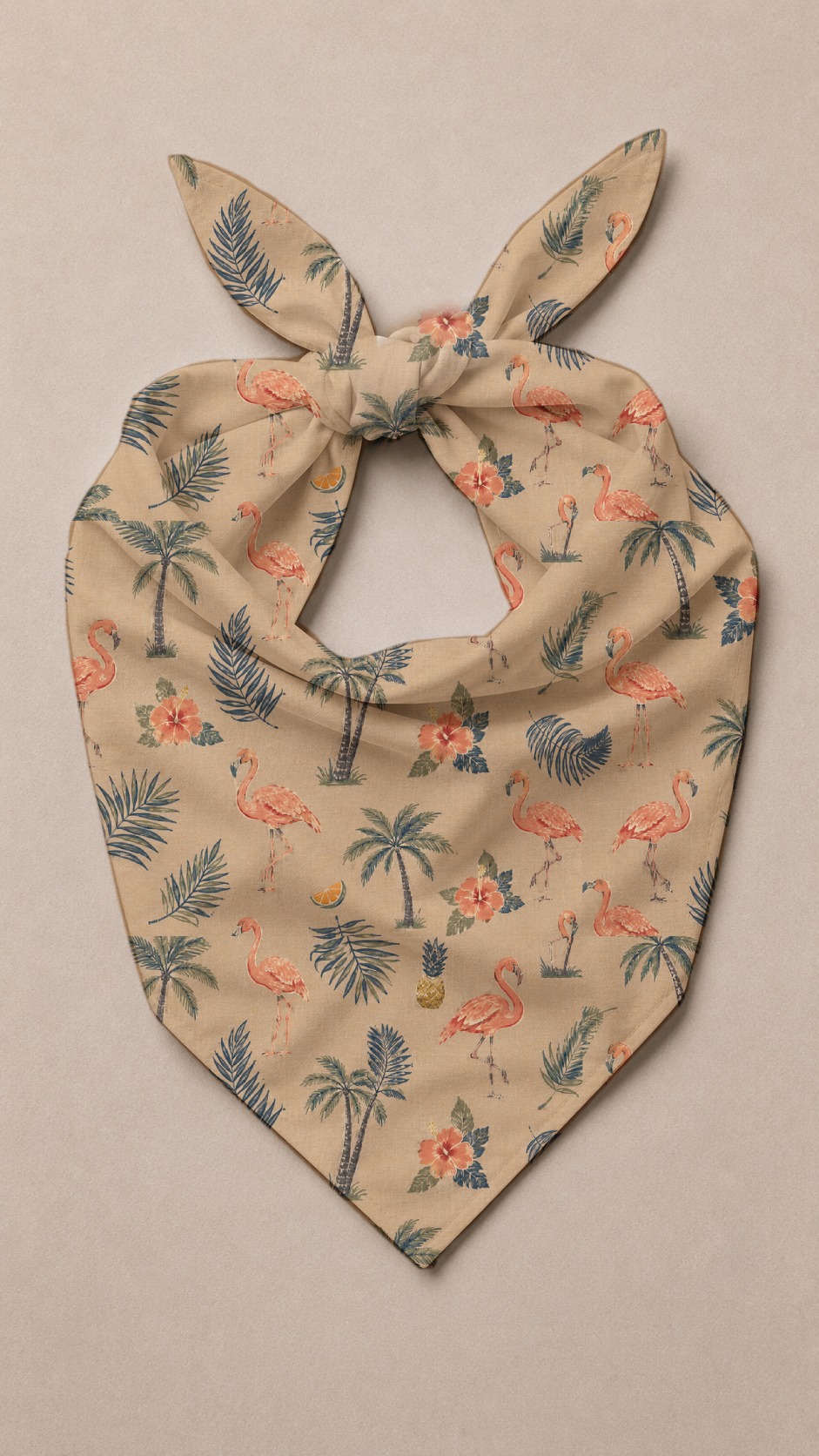 A beige bandana-style pet collar with a tropical print of pink flamingos, palm trees, and blue leaves, tied at the top and photographed flat on a neutral beige background.