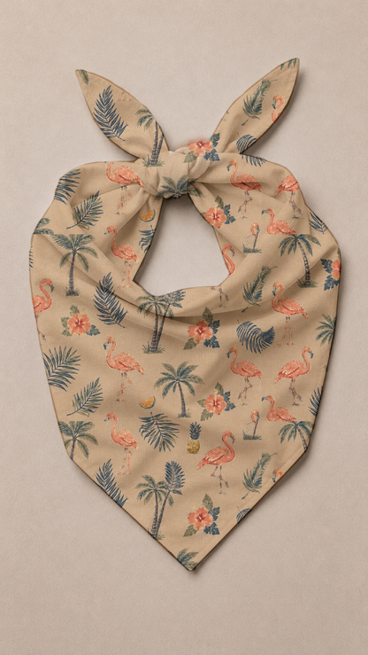 A beige bandana-style pet collar with a tropical print of pink flamingos, palm trees, and blue leaves, tied at the top and photographed flat on a neutral beige background.