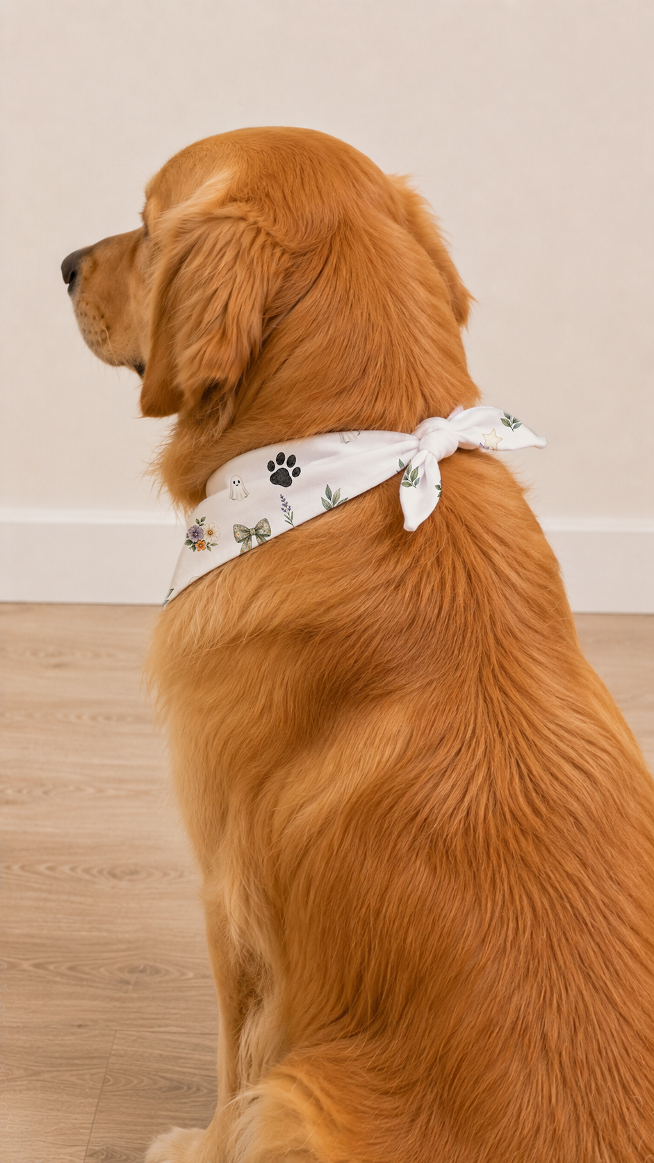 A golden retriever dog wearing a white bandana with a black paw print and green floral pattern, sitting on a light wood floor against a beige wall.