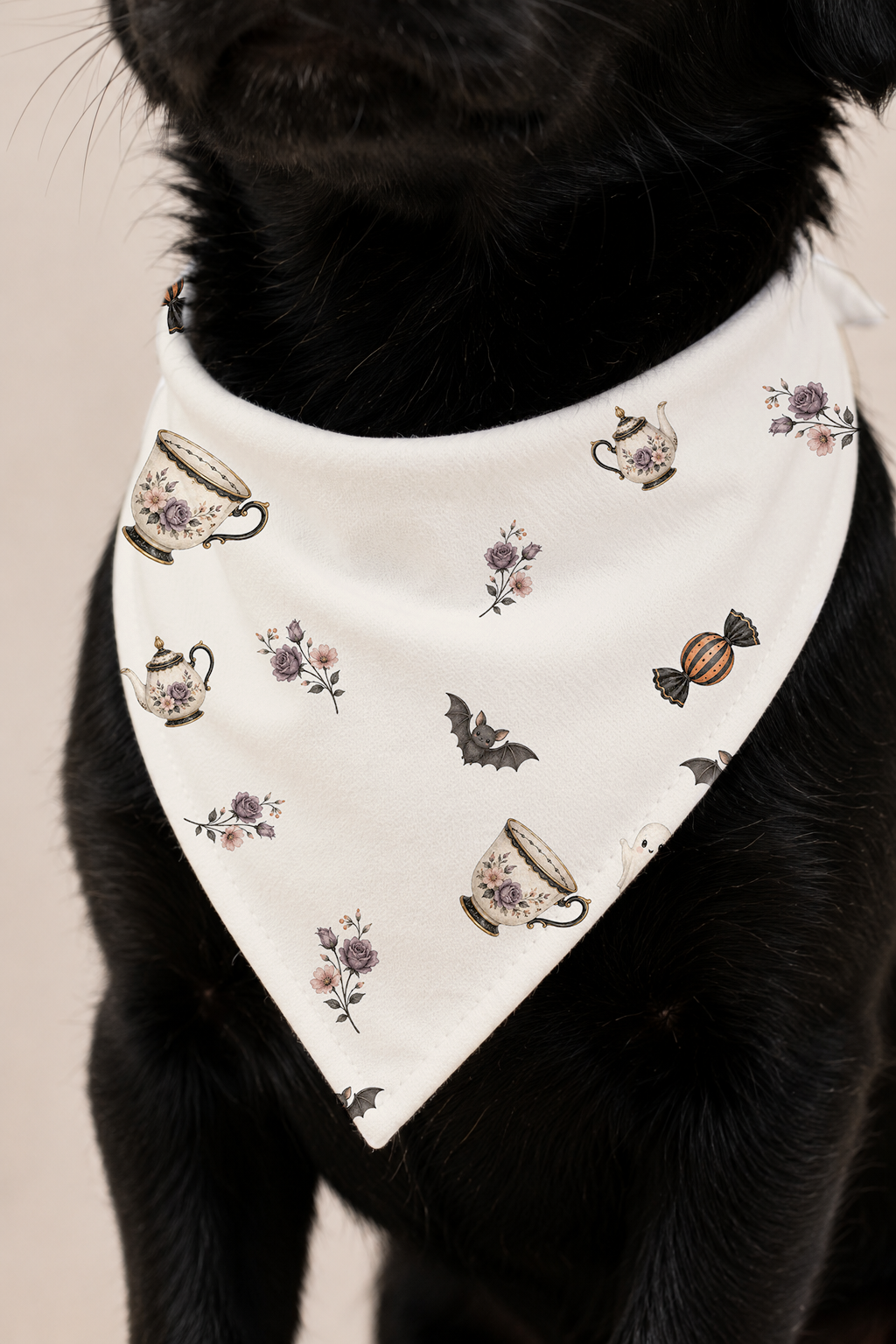 A close-up of a black dog wearing a white triangular bandana with a vintage tea-time pattern featuring teacups, bats, and candy.