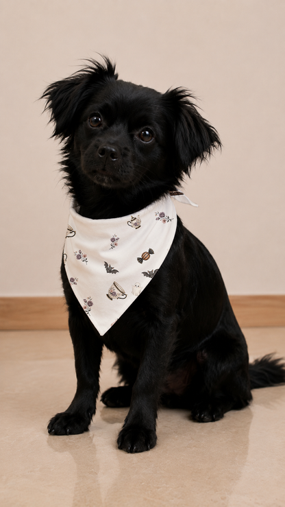 A small black dog wearing a white bandana with a cute teddy bear and bow tie pattern, sitting on a beige tiled floor against a beige wall.