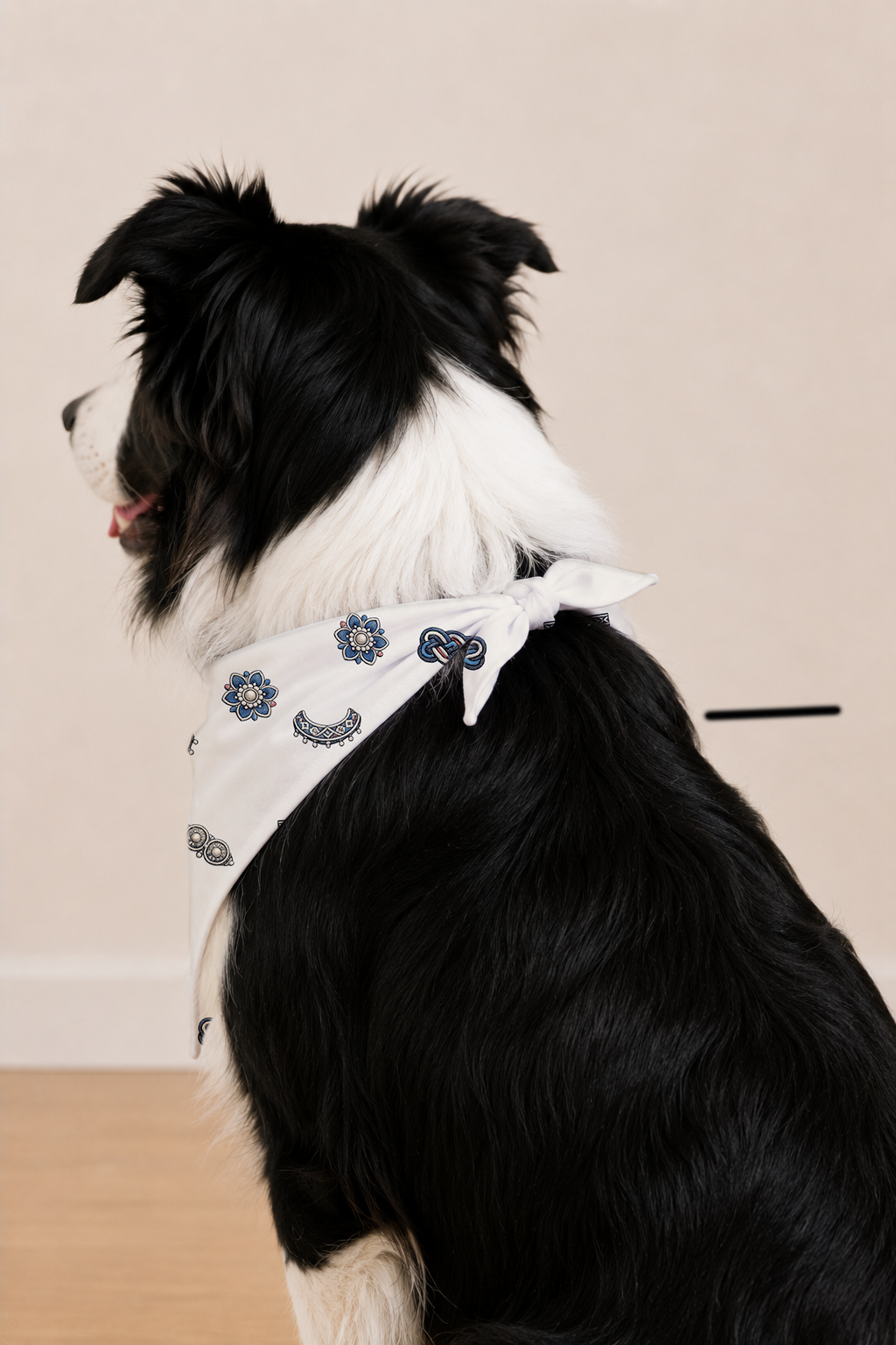 A black and white dog wearing a white bandana with blue paisley and floral patterns, shown from the side against a plain beige wall and light wood floor.