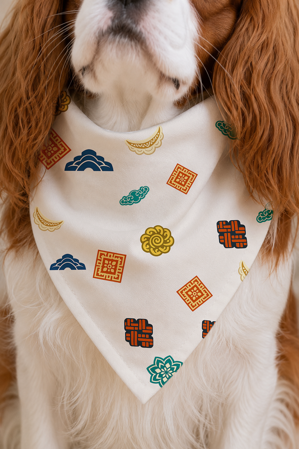 A close-up of a dog wearing a cream-colored bandana-style pet scarf with a colorful geometric and floral pattern.
