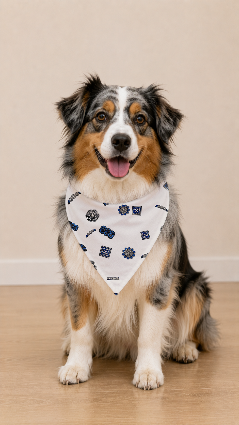 A smiling Australian Shepherd dog wearing a white triangular bandana with a navy blue gear pattern, sitting on a light wood floor against a beige wall.
