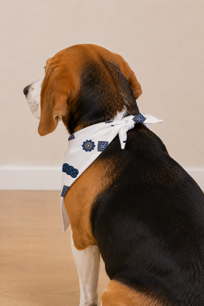 A beagle dog shown from the back and side, wearing a white bandana with blue patterns, sitting on a light wood floor against a beige wall.