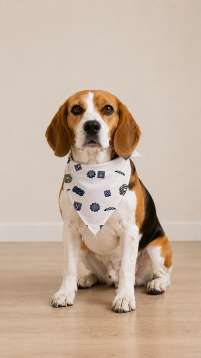 A beagle dog sitting on a light wood floor wearing a white bandana with a navy blue geometric pattern, set against a plain beige wall.