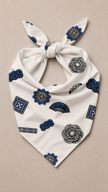 A white tie-front bandana or scarf featuring an all-over print of blue and gray decorative motifs, including flowers, geometric patterns, and fan shapes, laid flat on a light beige background.