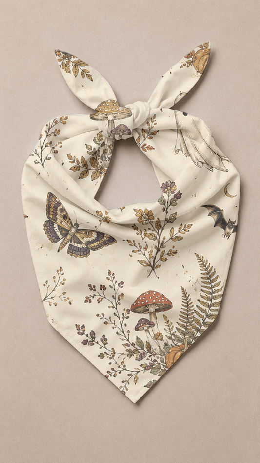 A cream-colored dog bandana with a knotted top, featuring a detailed print of woodland motifs including a butterfly, mushrooms, ferns, and leaves in earthy brown, beige, and muted purple tones, laid flat on a neutral beige background.