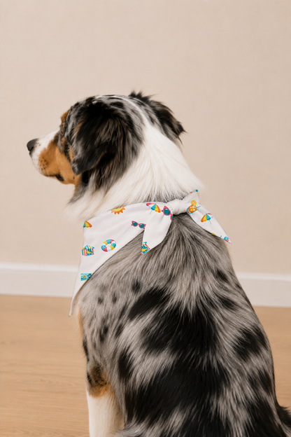 A dog wearing a white bandana with a colorful beach-themed print, photographed indoors against a plain beige wall and light wood floor.