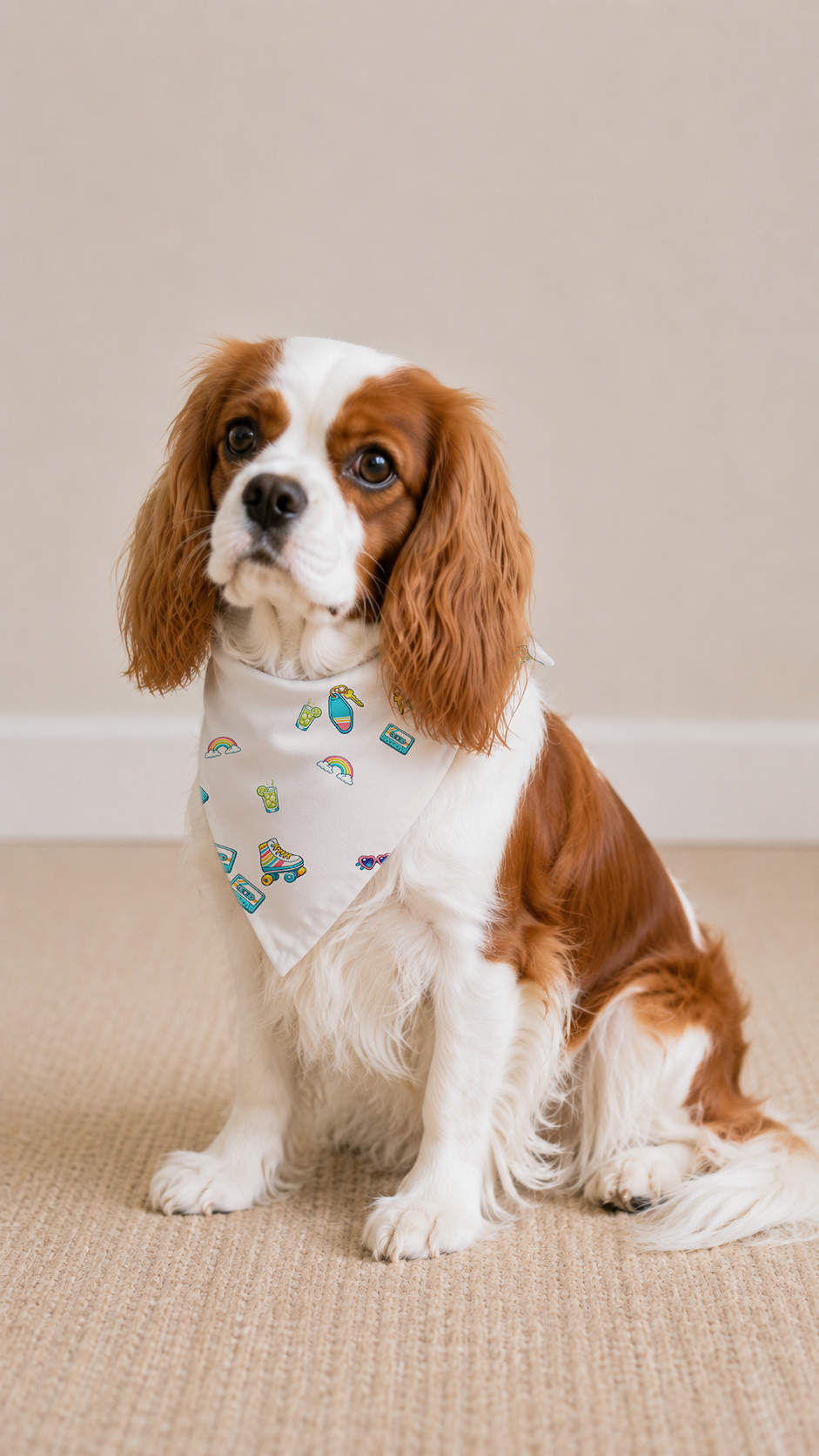 A brown and white Cavalier King Charles Spaniel dog wearing a white bandana with a colorful cartoon pattern, sitting on a beige textured rug against a neutral beige wall.