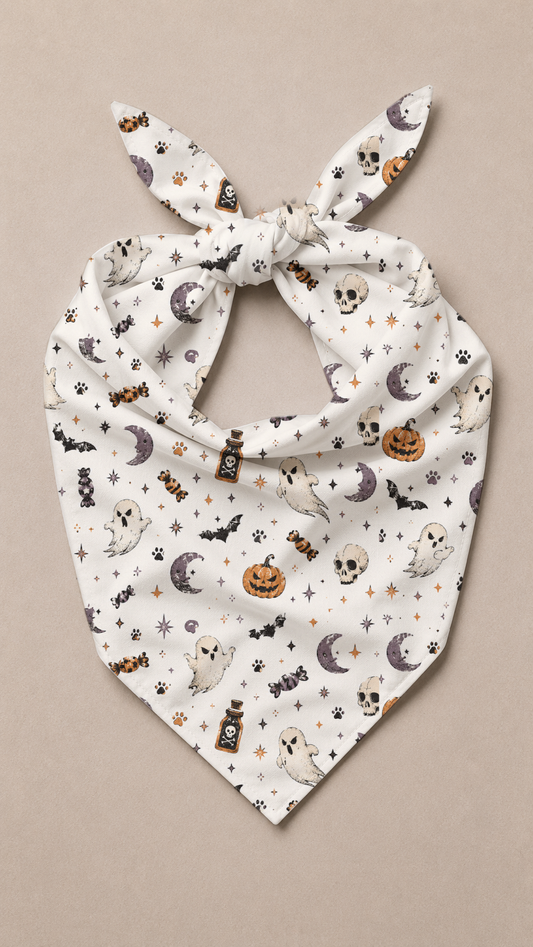 A white pet bandana with a knotted top and pointed bottom, featuring a repeating Halloween print of ghosts, pumpkins, bats, skulls, and moons, displayed flat against a neutral beige background.