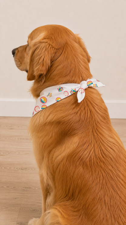 A golden retriever dog wearing a white pet bandana with a colorful beach print, sitting on a light wood floor against a neutral beige wall.