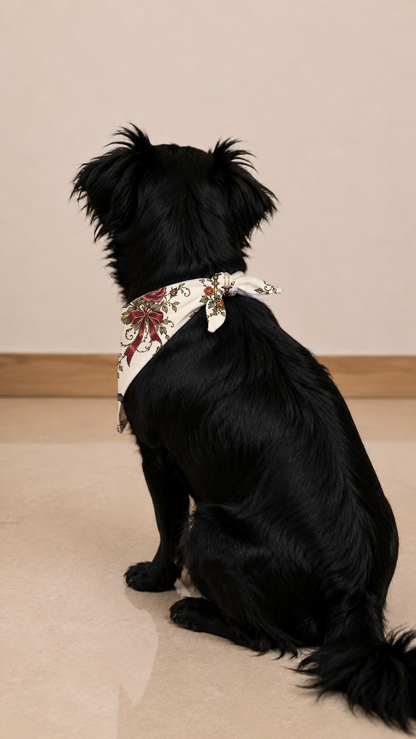 A black dog sitting on a beige tiled floor, wearing a white bandana with a red and green floral pattern.