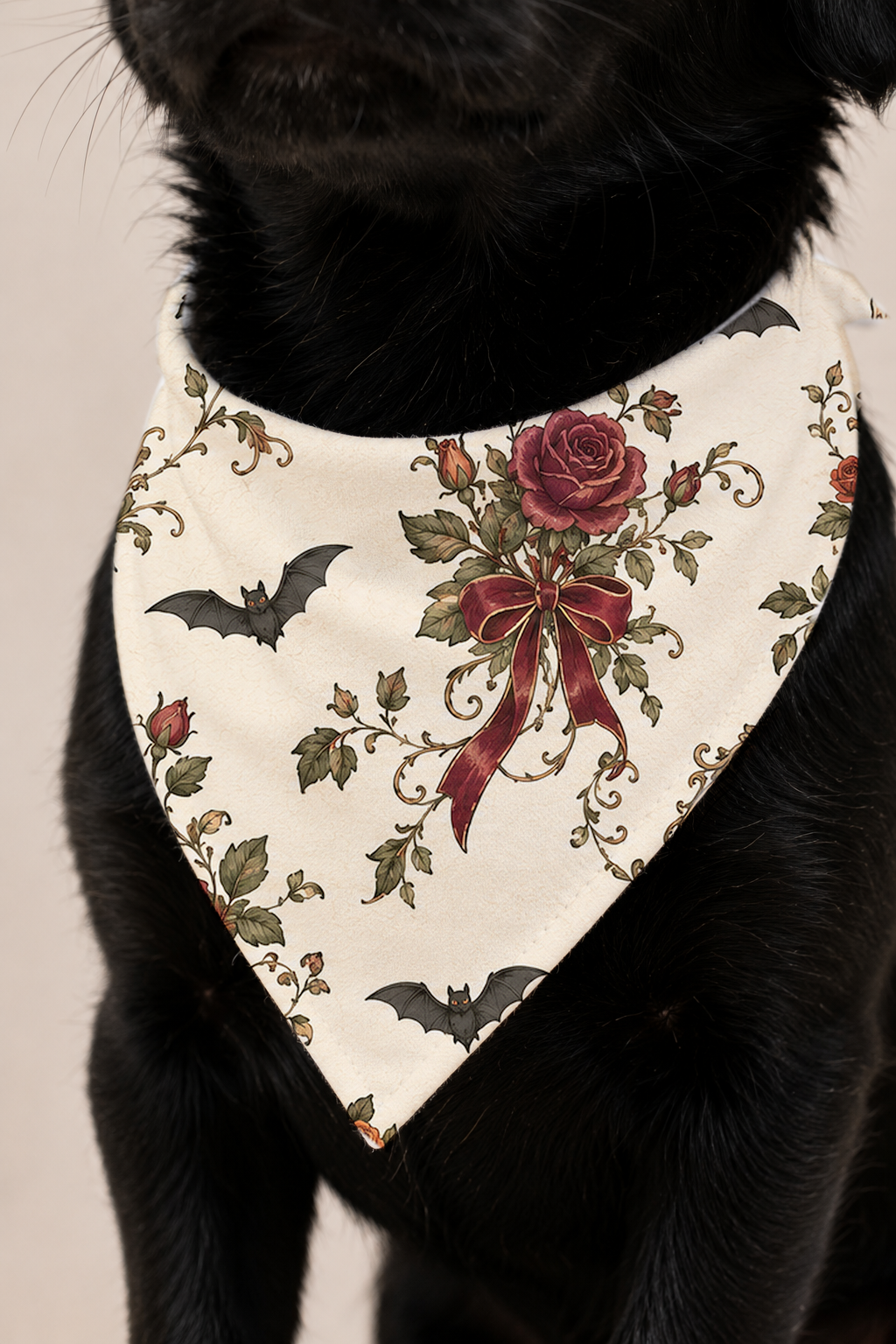 A black dog wearing a cream-colored bandana bib with a gothic design featuring red roses, a large red ribbon bow, green leaves, and black bats.